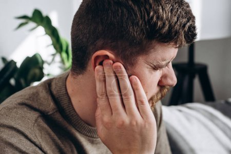 Photo for Close-up of unhealthy young caucasian man 30s touching ear, suffering from sudden throbbing ear ache, looking aside. Upset bearded male feeling unwell sitting on couch in living room at home - Royalty Free Image