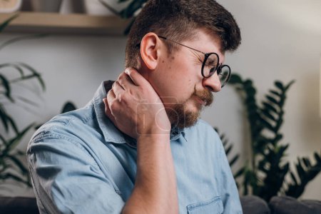 Photo for Young bearded man in eyeglasses studying until night at computer with neck pain. Close up of tired worker kneading neck, muscle recovery and massage. Neck pain, cervical osteochondrosis concept - Royalty Free Image