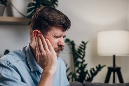Photo for Close-up of upset young guy 30s touches sore ears suffering from sudden throbbing earache. Unhealthy caucasian male feeling unwell sitting on grey couch in living room at home. Tired ears concept - Royalty Free Image