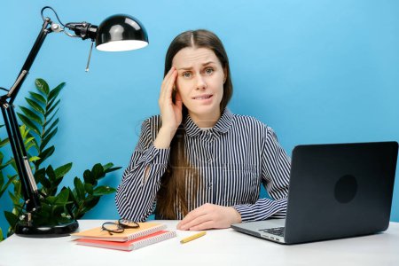 Foto de Retrato de la confundida secretaria decepcionada empleada mujer en camisa sentarse a trabajar en el escritorio de la oficina blanca con el ordenador portátil de PC apuntalar la frente, posando aislado sobre la pared de fondo de color azul en el estudio - Imagen libre de derechos