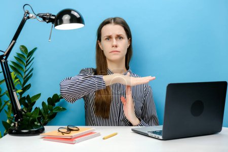 Foto de Estricta empleada joven mujer de negocios de 20 años en camisa sentado trabajo en el escritorio de la oficina blanca con el ordenador portátil que sostiene las manos de gesto de parada perpendicularmente, posando aislado sobre la pared de fondo de color azul en el estudio - Imagen libre de derechos