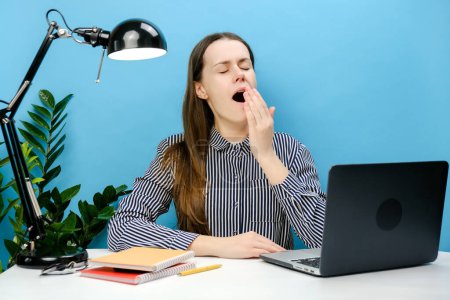 Foto de Retrato de cansada empleada agotada somnolienta joven mujer caucásica en camisa sentarse a trabajar en el escritorio de la oficina con ordenador portátil bostezo, posando aislado sobre pastel color azul fondo de la pared en el estudio - Imagen libre de derechos