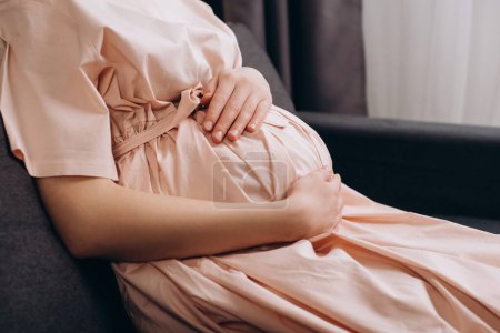 Photo for Close up of loving young pregnant woman sitting on couch stroking big belly, expressing love and care to unborn baby, expecting childbirth, feeling tenderness, resting alone at home. Maternity concept - Royalty Free Image
