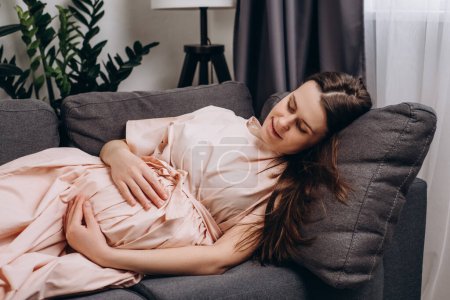 Téléchargez les photos : Adorable happy young pregnant woman in dress stroking belly, feeling unconditional love to unborn baby, expressing caress and devotion, enjoying carefree serene weekend lying on cozy couch at home - en image libre de droit