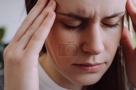 Téléchargez les photos : Close up portrait of displeased young woman suffering from migraine while staying alone at apartment. Unhappy millennial lady massaging temples, mental stress, panic attack touching aching sore head - en image libre de droit