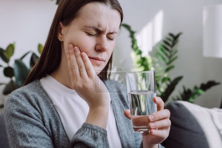 Téléchargez les photos : Portrait of young woman feel terrible toothache after drink cold water. Female sitting on couch at home touching cheek, feel hurt and suffering from sensitive tooth ache. Pain and cavities concept - en image libre de droit