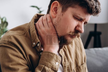 Foto de Close up of unhappy caucasian male holding hand on neck, suffering from neck pain, incorrect posture, joint inflammation. Unhealthy sad bearded man sitting on couch needs medicine chiropractor help - Imagen libre de derechos
