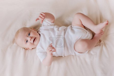 Draufsicht von glücklichen liebenswerten kleinen neugeborenen Jungen mit lächelndem Gesicht auf gemütlichem Bett im Wohnzimmer zu Hause liegend. Fröhliches, hübsches Baby, das auf einer bequemen Decke liegend spielt. Kindheitskonzept
