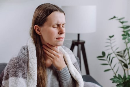 Symptoms from flu season in winter concept. Close-up of unhealthy sad young woman covered blanket sitting on grey couch at home suffering from sore throat, angina, hard to swallow, voice loss