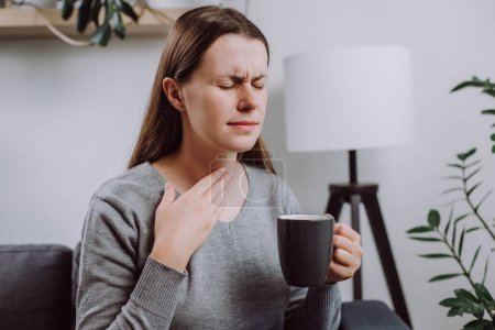 Photo for Sick unhappy young caucasian female 25s touch neck, have sore throat, holding mug, drink warm water, have a fever, flu in weakness, sitting alone on couch at home. Health care on virus seasonal - Royalty Free Image