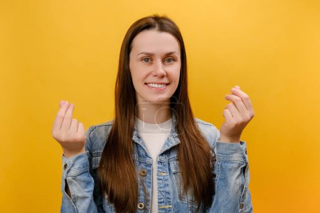 Photo for Portrait of attractive smiling young woman 25s looking at camera with money or italian gesture with hands, wearing denim jacket, posing isolated over plain yellow color background wall in studio - Royalty Free Image