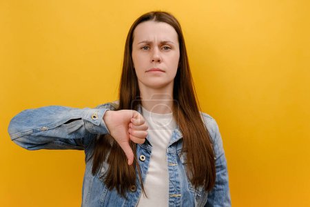 Portrait of upset dissatisfied young woman showing thumb down and frowning face in disgust, negative feedback, wearing denim jacket, posing isolated over yellow color background wall in studio