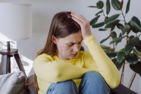 Upset young woman frustrated by problem with work or relationships, sit on couch at home, embracing knees, covered face in hand, feeling despair and anxiety, loneliness, having psychological trouble