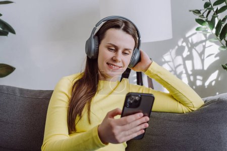 Portrait of happy peaceful young caucasian woman hold phone relaxing on comfortable couch, listening to tranquil classical music in wireless headphones, enjoying lazy weekend hobby time alone at home