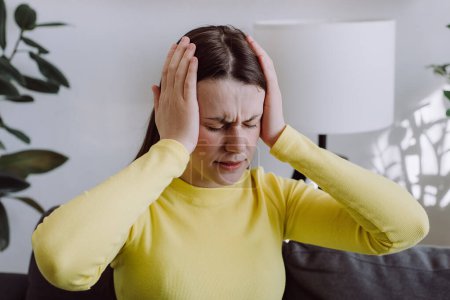 Worried sad young caucasian female has dizziness or headache because migraine sitting alone on couch in living room at home. Brain stroke or low blood pressure symptom on people medical concept