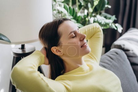 Téléchargez les photos : Gros plan d'une jeune femme paisible souriante aux yeux fermés se relaxant sur un canapé confortable à la maison, calme et attentionnée penchant vers l'arrière, visualisant un bon avenir, faisant la sieste ou rêvant, profitant du temps libre - en image libre de droit