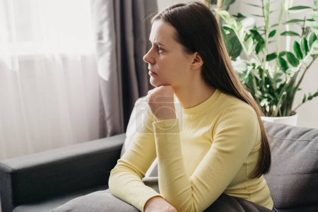 Anxious worried young woman sitting on sofa at home. Frustrated confused caucasian female feels unhappy, problems in personal life, quarrel break up with boyfriend and unexpected pregnancy concept