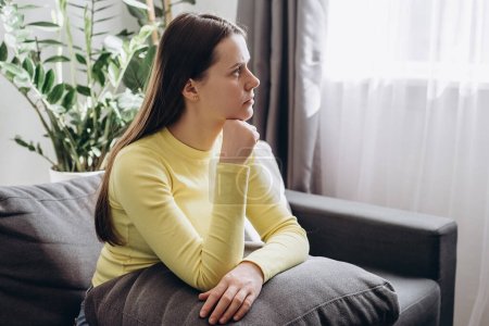 Umsichtige junge Frau, die zu Hause auf der Couch sitzt, blickt in die Ferne und trauert, unglücklich aufgewühlte Mädchen denken an Beziehungsprobleme, kämpfen mit Fehlgeburten oder Abtreibungsschwierigkeiten