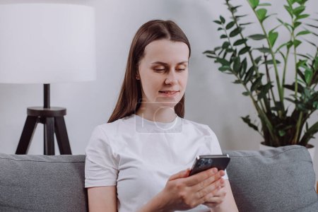 Cute young woman using cellphone sitting on cozy sofa at home, chatting via social media or messenger, enjoy new mobile app for fun, do shopping through retail services spend carefree pastime indoors