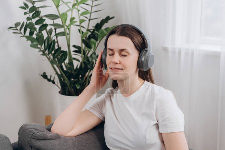 Carefree attractive smiling young woman sitting on cozy sofa in headphones enjoy favorite music or listen audiobook, spend weekend time alone at home after hard work day. Time to relax, chill out