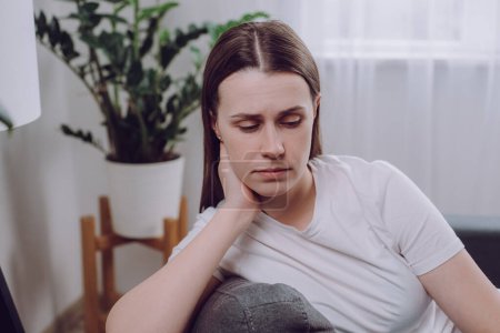 Confused young woman feels unhappy, problems in personal life, quarrel break up with boyfriend and unexpected pregnancy concept. Anxious worried caucasian female sitting alone on sofa at home