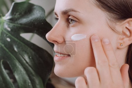 Close up of smiling pretty young caucasian woman applies face cream, applying cream in motions, skin care, natural cosmetics, face care concept