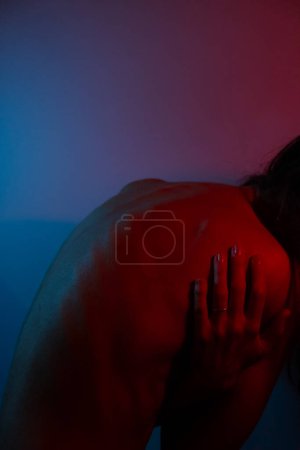 Photo for Intense young woman, blue and red lighting on back. Neck and back pain.Mental burden concept.Bent woman with bare back - Royalty Free Image