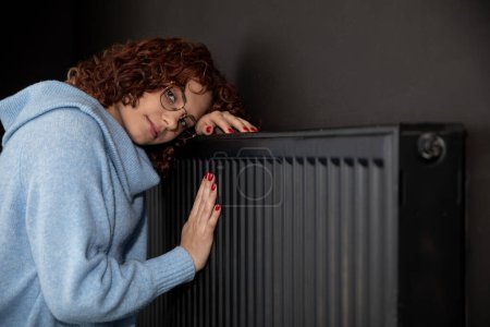 Téléchargez les photos : Une jeune fille dans un pull épais bleu étreint un radiateur chaud. Chauffage central nécessaire dans la maison pendant la période d'hiver. - en image libre de droit