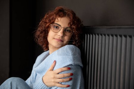 Foto de Una chica con un suéter azul grueso se sienta junto al radiador. Calefacción central de mal funcionamiento. Una sonrisa suave. - Imagen libre de derechos