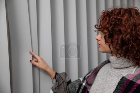 A young girl with curly hair and wearing glasses gently unveils the vertical blinds and looks through the ok no outside.