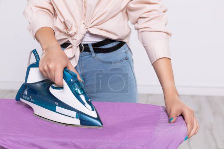 Téléchargez les photos : A woman irons a purple mens shirt with an iron on an ironing board. Close-up view. A womans hand with nails in blue with glitter. - en image libre de droit