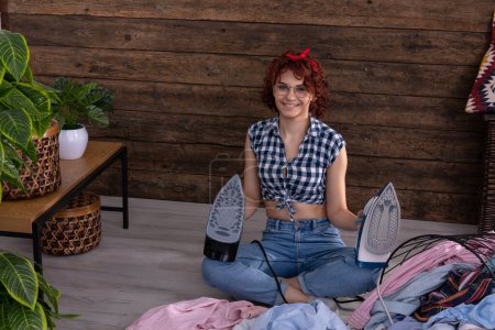 Photo for An attractive girl sits on the floor of the house. A young woman with short and curly hair. She holds an iron in her hand. A wall of brown boards. - Royalty Free Image