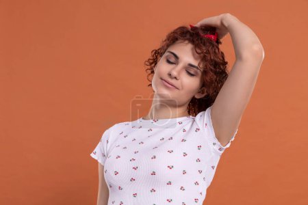 Photo for Curly chestnut-colored hair. A woman corrects her hair gently with her hand. White blouse with a cherry pattern. Isolated from the uniform background. - Royalty Free Image