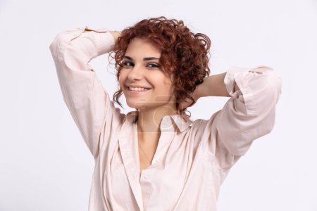 Téléchargez les photos : Smiling and full of sincere joy, the girl raises her hair up with her hands. Beautiful short curly chestnut-colored hair. Natural hairstyle. - en image libre de droit