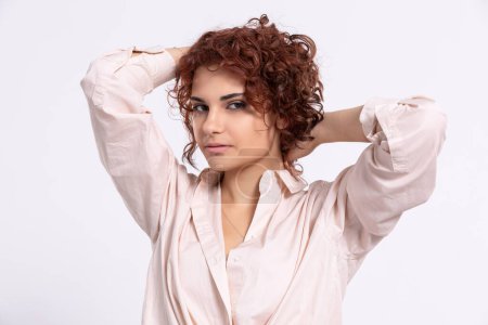 Téléchargez les photos : The girl raises her hair up with her hands. Beautiful short curly chestnut-colored hair. Natural hairstyle. Light-colored long-sleeved blouse. - en image libre de droit