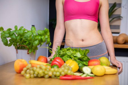 Photo for Lots of vegetables and fruits lying on the kitchen table. Behind the table a silhouette of a woman with an exposed belly in a tight pink sports top leaning against the table top - Royalty Free Image