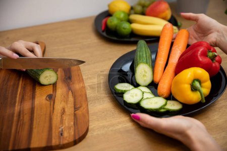Foto de Autumn colored vegetables cut by a woman on a brown board. She slices peppers and carrots with a sharp knife. - Imagen libre de derechos