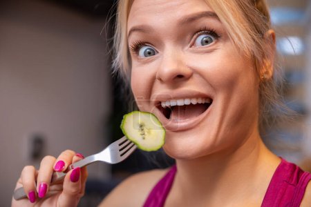 Foto de Large close-up on the face of a blonde woman with her mouth open. The woman holds a fork with a scooped cucumber slice with skin and brings it close to her open mouth. - Imagen libre de derechos