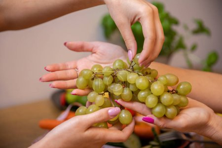 Foto de Las manos femeninas sostienen un ramo de uvas verdes. Manos de dos mujeres con uñas de colores. Gran primer plano en dos pares de manos. Hierbas borrosas en una olla en el fondo. - Imagen libre de derechos
