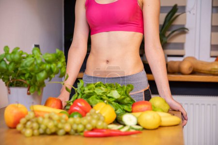 Photo for Lots of vegetables and fruits lying on the kitchen table. Behind the table a silhouette of a woman with an exposed belly in a tight pink sports top leaning against the table top - Royalty Free Image