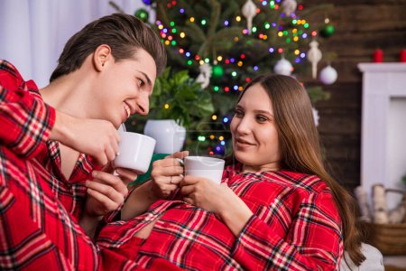 Photo for A couple expecting a baby lies on a gray couch against a backdrop of a dressed Christmas tree with colorful lights. The man and woman are wearing identical pajamas and holding white teacups. - Royalty Free Image