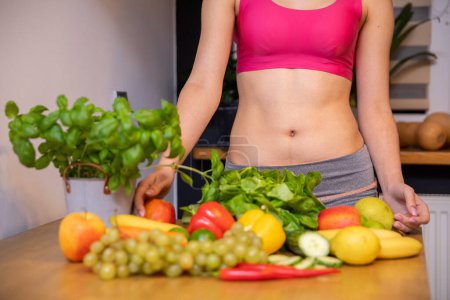 Photo for An athletic woman stands behind a table on which vegetables, fruits and herbs lie. She is dressed in a short sports tight top and only her torso is visible. Her muscular belly is exposed. - Royalty Free Image