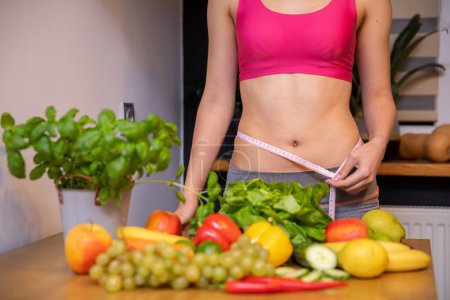 Photo for An athletic woman with a narrow waist wraps measure tape around her waist. In front of the womans figure stands a table and on it lie colorful vegetables, fruits and herbs. In the background is a - Royalty Free Image