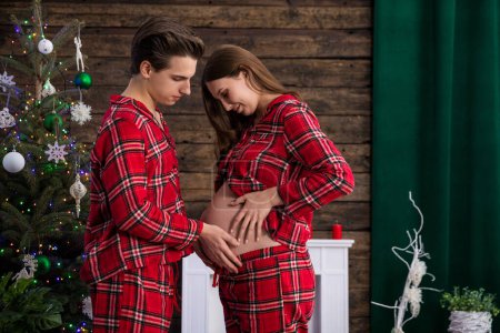 Photo for An expectant couple faces each other in a room with Christmas decorations and a Christmas tree. The man and the woman hold their hands on the pregnant belly. - Royalty Free Image