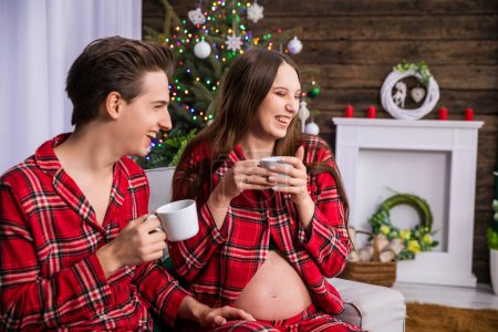 Photo for A woman in advanced pregnancy sits next to her partner on a gray couch. A fuzzy Christmas tree and Christmas decorations can be seen in the background. The couple is laughing and holding white teacups - Royalty Free Image
