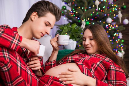 Photo for A couple expecting a baby lies on a couch against a backdrop of a dressed Christmas tree. The man and woman are holding white teacups. The woman has placed her hand on her pregnant belly. - Royalty Free Image