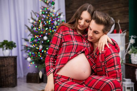 Photo for A man looks down at his partners pregnant belly while holding her in his lap. The couple is wearing identical red checkered pajamas. A smudged Christmas tree with lights stands in the background. - Royalty Free Image