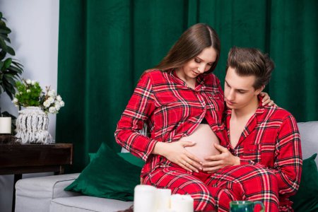 Photo for Girl sits on the lap of her boyfriend, who touches her pregnant bellyAn expecting couple sits on a gray sofa. The man touches his partners pregnant belly. In the background you can see a velvet heavy - Royalty Free Image