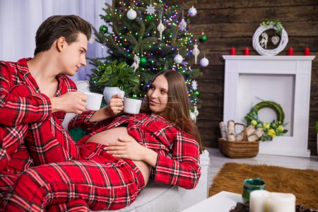 Photo for A man sitting on a couch holds the legs of a woman lying next to him on his lap. The woman is advanced in pregnancy and has an exposed belly. Both are holding white cups. - Royalty Free Image