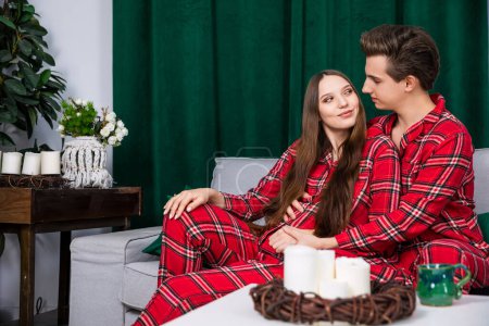 Foto de The couple in the photo shoot pose in identical red checkered pajamas. A heavy green curtain is seen in the background. The setting is romantic and in trendy colors of gray and green. - Imagen libre de derechos